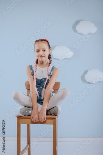 Stylish teenage girl is enjoying her modern kanekalon hair. little girl in fashionable denim overalls.girl sits on a wooden chair in the studio