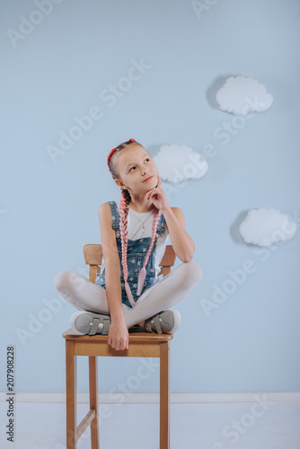 Stylish teenage girl is enjoying her modern kanekalon hair. little girl in fashionable denim overalls.girl sits on a wooden chair in the studio