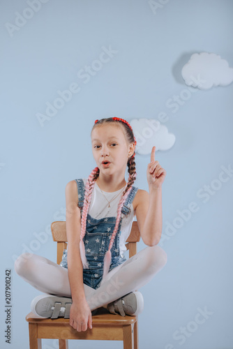 Stylish teenage girl is enjoying her modern kanekalon hair. little girl in fashionable denim overalls.girl sits on a wooden chair in the studio