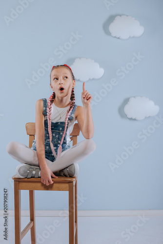 Stylish teenage girl is enjoying her modern kanekalon hair. little girl in fashionable denim overalls.girl sits on a wooden chair in the studio