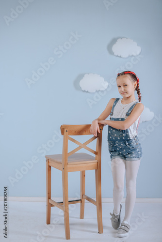 Stylish teenage girl is enjoying her modern kanekalon hair. little girl in fashionable denim overalls.girl sits on a wooden chair in the studio