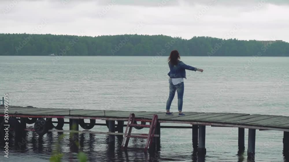 Woman in blue clothes walks on old wooden pier looks at camera then turns around and shows ahead with hand Woman in blue clothes walks on old wooden pier looks at camera then turns around and shows ahead with hand