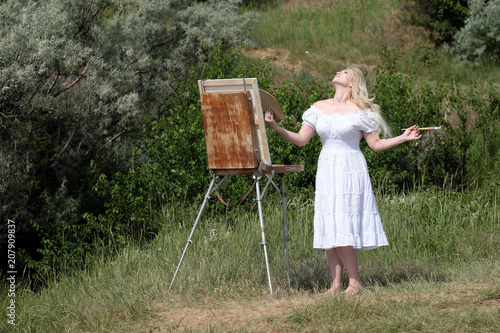 Beautiful girl draws a picture in the park using a palette with paints. Easel and canvas with a picture. photo