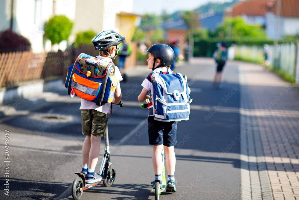 Two school kid boys in safety helmet riding with scooter in the city with backpack on sunny day. Happy children in colorful clothes biking on way to school.