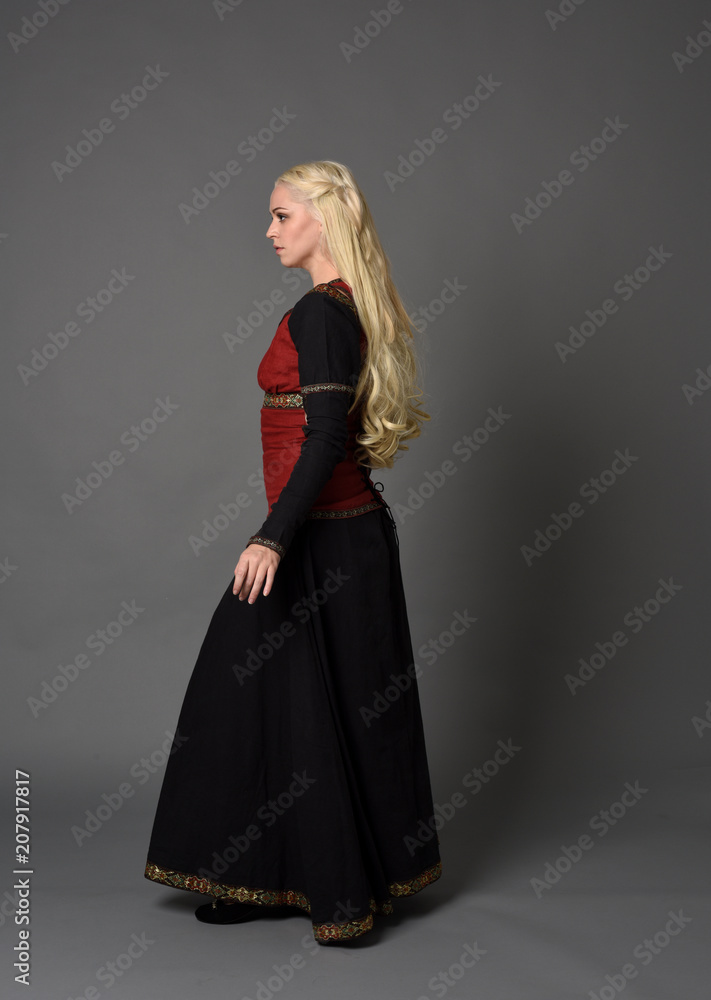 full length portrait of pretty blonde lady wearing a red and black fantasy medieval gown. standing pose on grey background. full length portrait of pretty blonde lady wearing a red and black fantasy medieval gown. standing pose on grey background.