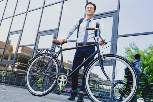 low angle view of handsome young businessman in eyeglasses standing with bicycle near modern building