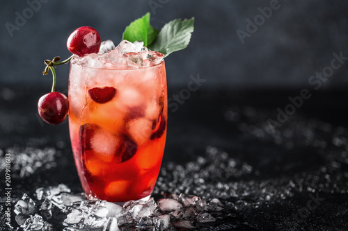 Fresh cherry cocktail. Fresh summer cocktail with cherry and ice cubes. Glass of cherry soda drink on dark stone background.