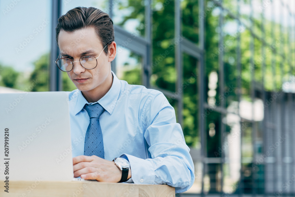 handsome young businessman in eyeglasses using laptop outside modern building handsome young businessman in eyeglasses using laptop outside modern building