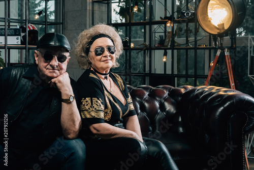 Unusual retired couple.Happy couple of retirees in biker clothes.pair of seniors in stylish black clothes sitting on a leather couch.Senior man in black leather jacket and goggles