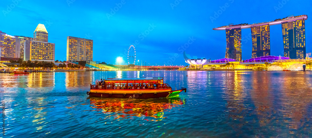 Fototapeta premium Panorama Singapuru budynków, drapaczy chmur i diabelskiego młyna odzwierciedlona w morzu. Statek turystyczny pływa w zatoce wieczorem. Panoramę Singapuru o niebieskiej godzinie. Nocna scena nabrzeża marina bay.