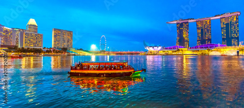 Panorama of Singapore buildings, skyscrapers and ferris wheel reflected in the sea. Tourist boat sails in the bay at evening. Singapore skyline at blue hour. Night scene waterfront marina bay.