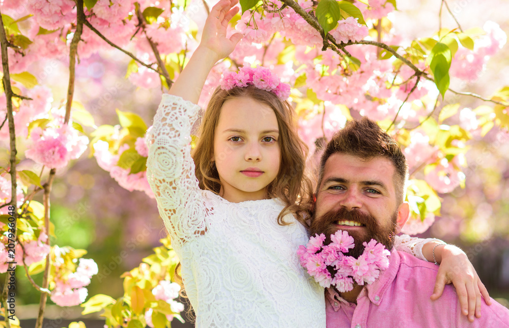 Fototapeta premium Girlish leisure concept. Girl with dad near sakura flowers on spring day. Child and man with tender pink flowers in beard, wreath on head. Father and daughter on happy faces hugs, sakura background.