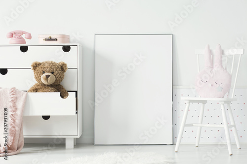 Mockup of empty poster between cabinet with teddy bear and white chair in kid's interior. Real photo. Paste your poster here
