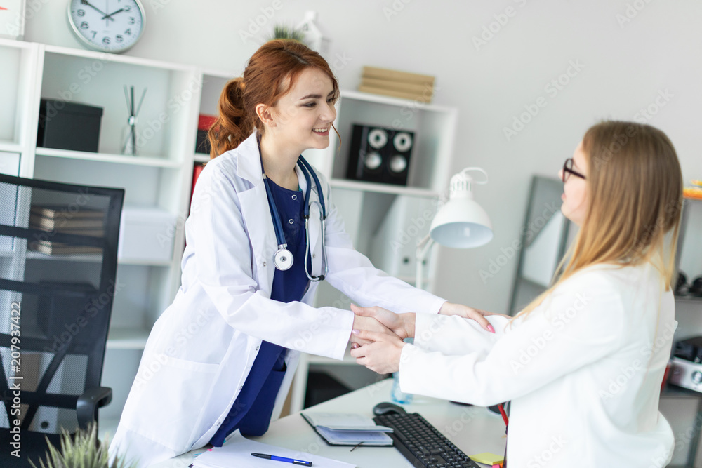 A beautiful young girl in a white coat is standing near the desk in the office and shaking hands with the patient. The patient thanks the doctor. A beautiful young girl in a white coat is standing near the desk in the office and shaking hands with the patient. The patient thanks the doctor.