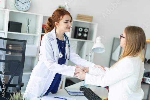 Fototapeta Naklejka Na Ścianę i Meble -  A beautiful young girl in a white coat is standing near the desk in the office and shaking hands with the patient. The patient thanks the doctor.