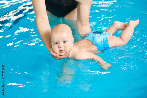 Little baby swimming in water pool with help from mothers hands.