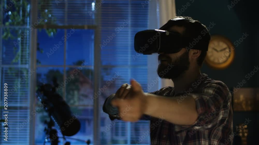Casual man sitting on bed wearing VR goggles and touching air around having fun Casual man sitting on bed wearing VR goggles and touching air around having fun