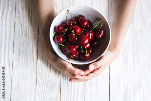 delicious ripe cherry in a bowl in children's hands on white wooden background photo