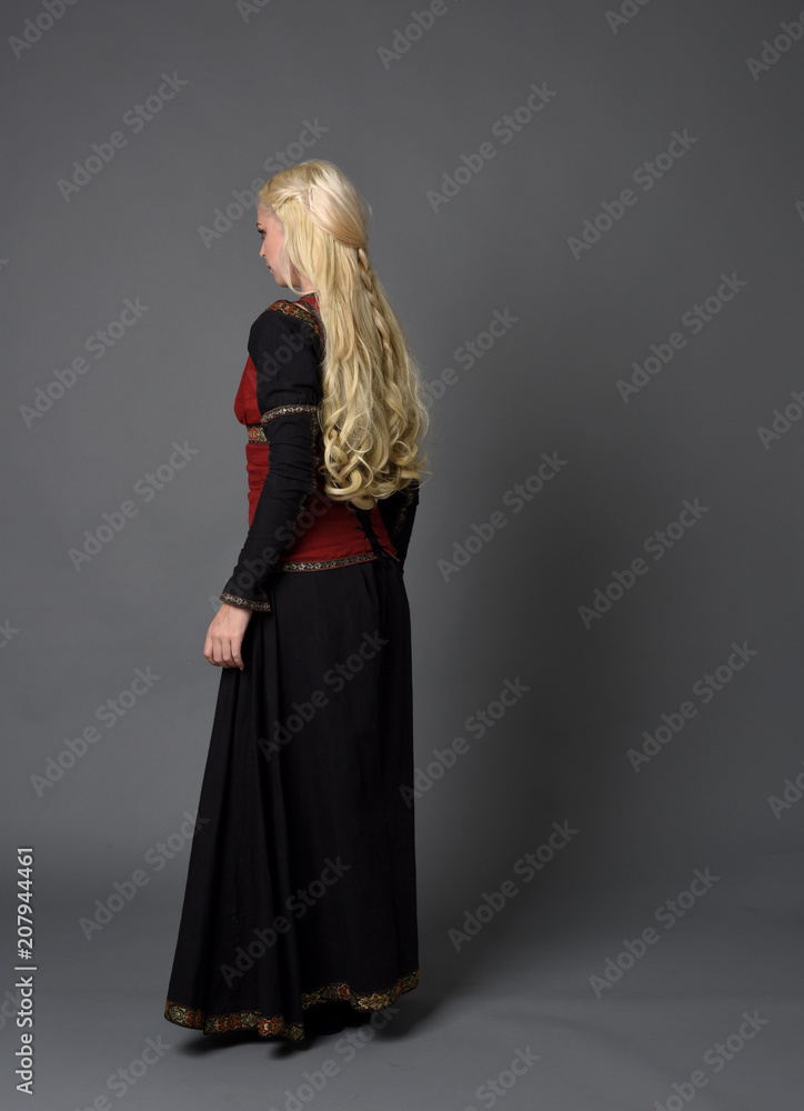 Fototapeta premium full length portrait of pretty blonde lady wearing a red and black fantasy medieval gown. standing pose on grey background.