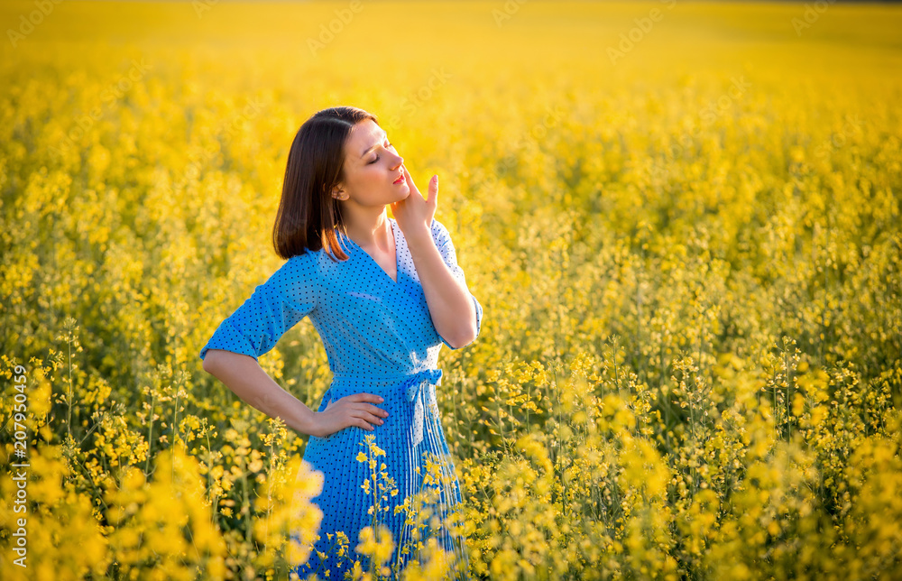 Allergy free concept. Freedom. Attractive girl in blue dress on yellow blooming meadow. Summer sunny portrait of brunette girl. Allergy free concept. Freedom. Attractive girl in blue dress on yellow blooming meadow. Summer sunny portrait of brunette girl.