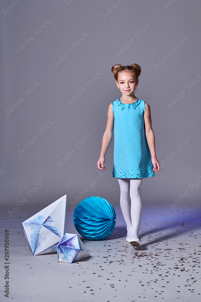 Elegant fashion child girl model goes in bright blue dress.Designer  collection. Beautiful romantic lady. Teenage girl, poses for children's  clothing catalog in studio on gray background. Stock Photo