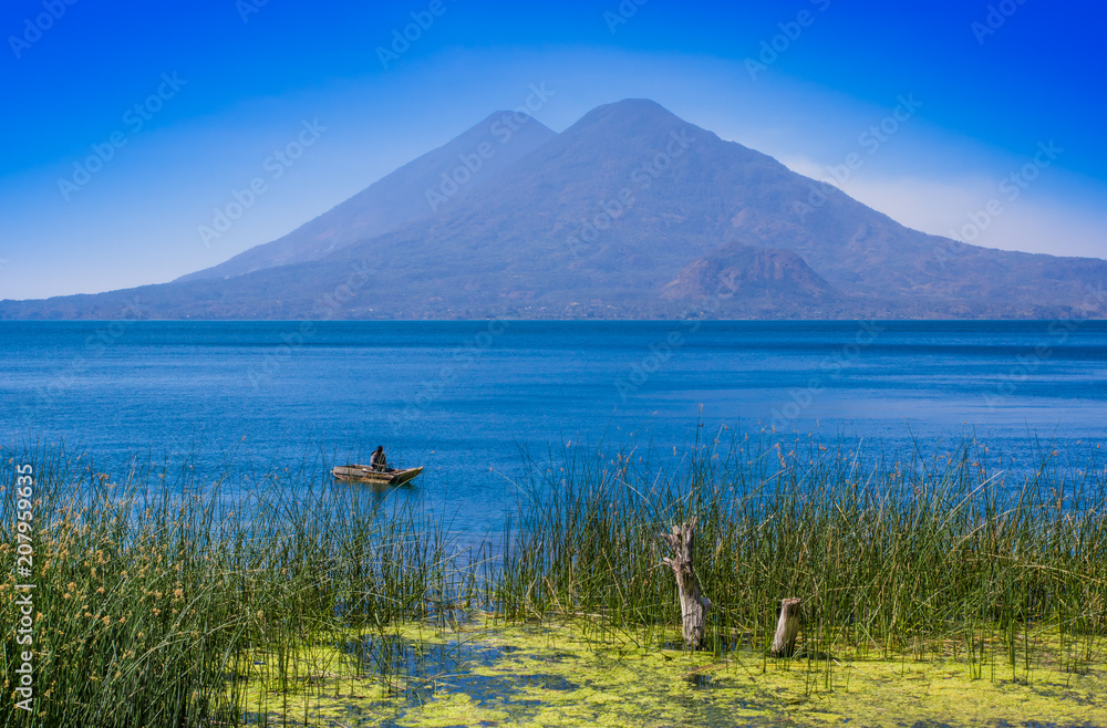 Fototapeta premium Outdoor view of totora acuatic plants in the border of the lake, with unidentified fisherman in a small boats in Atitlan Lake, with San Pedro volcano in Background