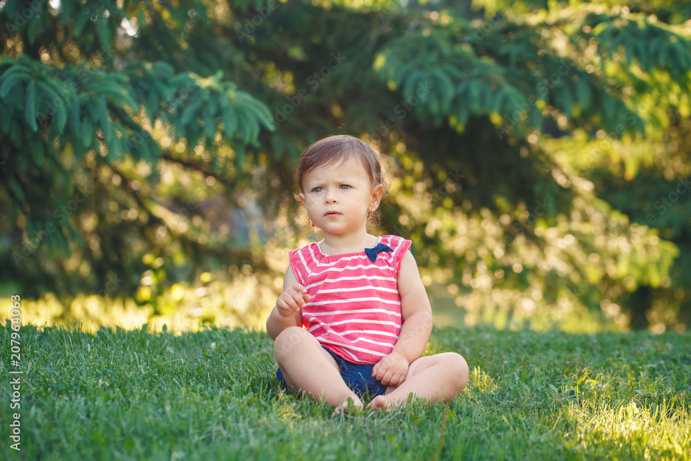 Portrait of cute adorable pensive serious little Caucasian baby girl toddler in park outside. Happy lifestyle childhood concept. Child kid sitting on grass during summer sunset Portrait of cute adorable pensive serious little Caucasian baby girl toddler in park outside. Happy lifestyle childhood concept. Child kid sitting on grass during summer sunset