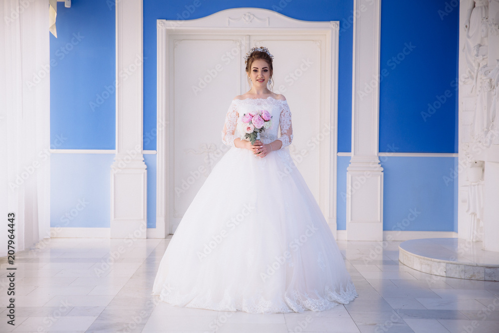 A beautiful girl in a lush white dress on her wedding day is standing with a bouquet of peonies in a blue hall with a high ceiling