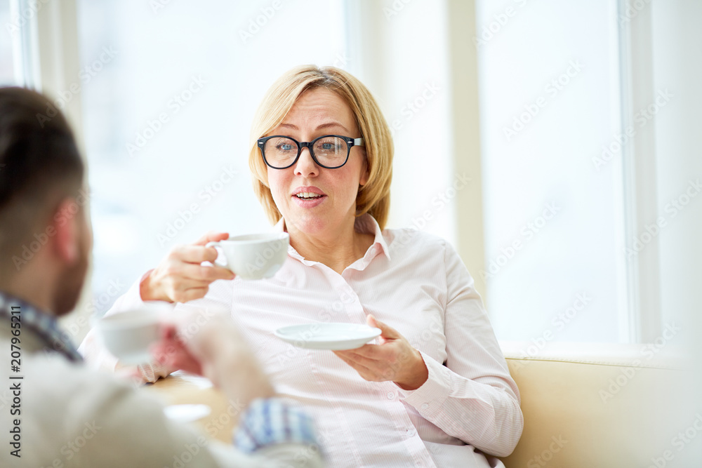 Adult woman in glasses sitting on couch with man enjoying coffee and talking in leisure. Adult woman in glasses sitting on couch with man enjoying coffee and talking in leisure.