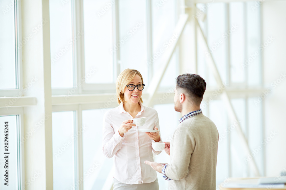Adult woman speaking to young man while having coffee and standing in light spacious room. Adult woman speaking to young man while having coffee and standing in light spacious room.