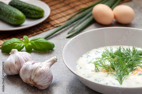White plate with cucumber cold soup with fresh dill on a concrete countertop with garlic, cucumbers, eggs and chives