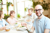 Crop view of smiling bald male in glasses holding glass with water sitting with friends in cafe and looking at camera