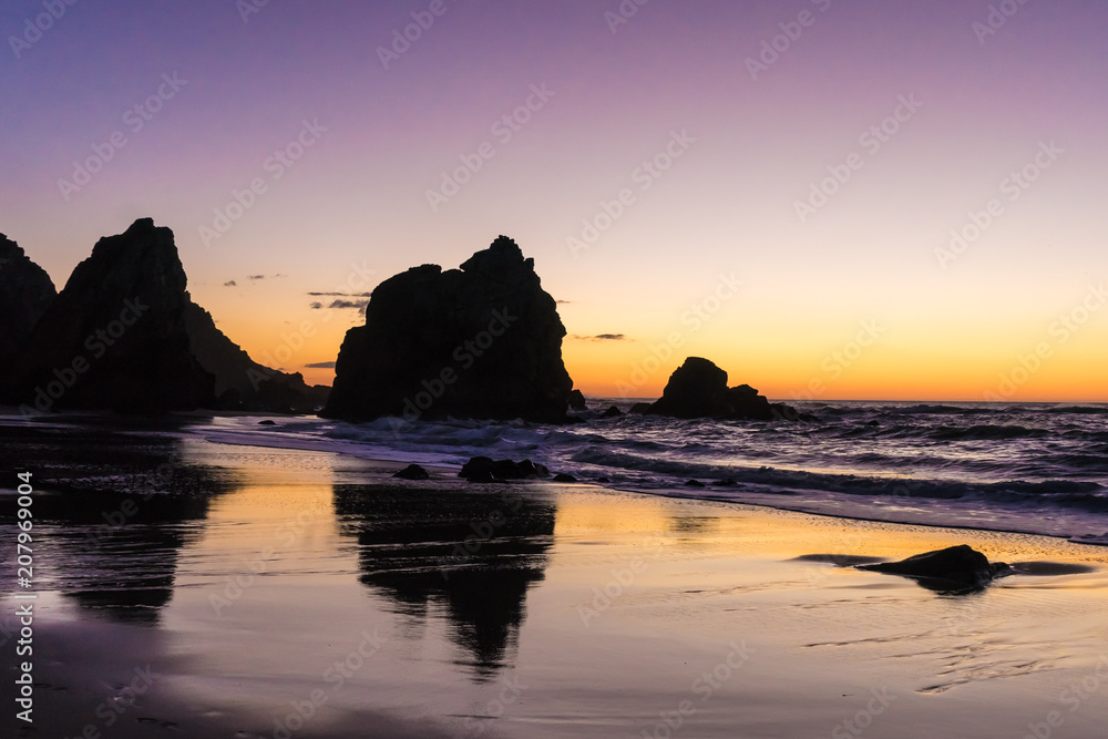 Reflections of giant rocks in the wet sand at sunset at the Praia da Ursa, Portugal