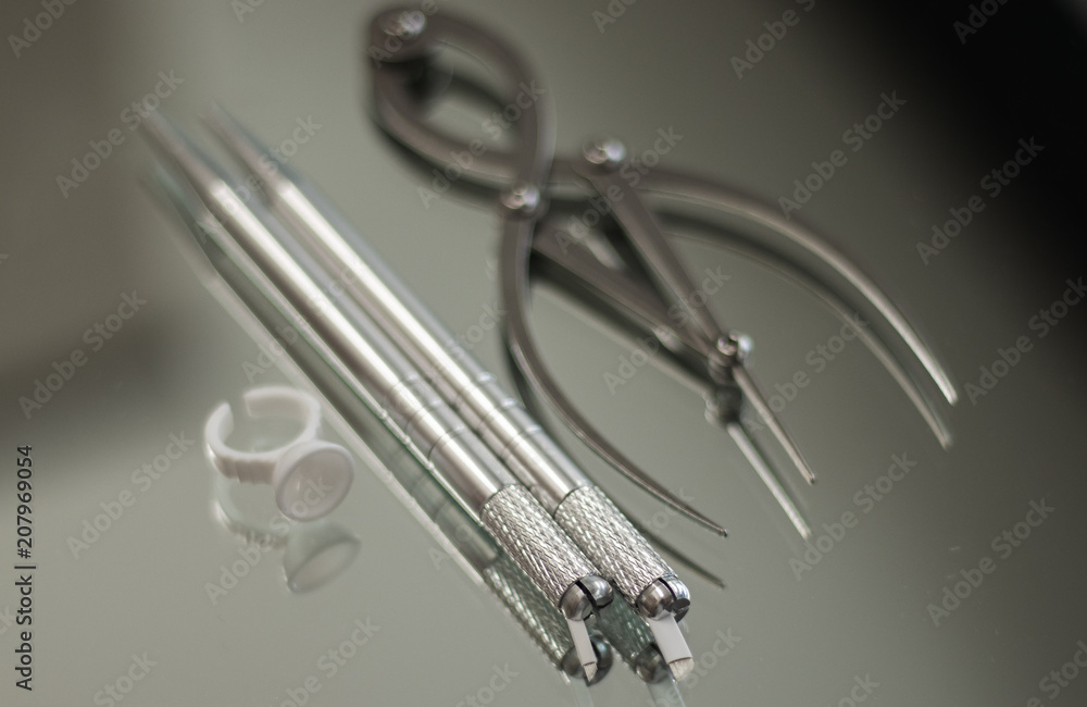 An ink ring, microblading pen, and brow caliper on a mirror surface An ink ring, microblading pen, and brow caliper on a mirror surface