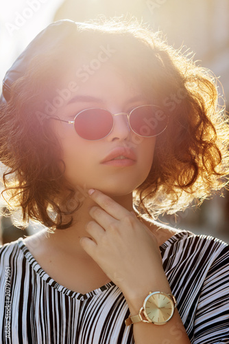 Young beautiful stylish curly woman wearing oval red color sunglasses, golden wrist watch on the hand, striped dress. Model posing in street. Sunny day natural light. Summer fashion concept
