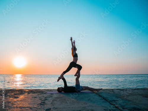 Fit young couple doing acro-yoga at sea beach. Man lying on concrete plates and balancing woman on his feet. Beautiful pair practicing yoga together.