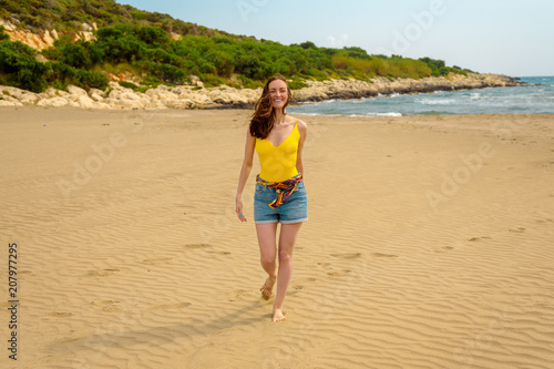 A woman in a yellow swimsuit and denim shorts walking barefoot on the sand on the beach