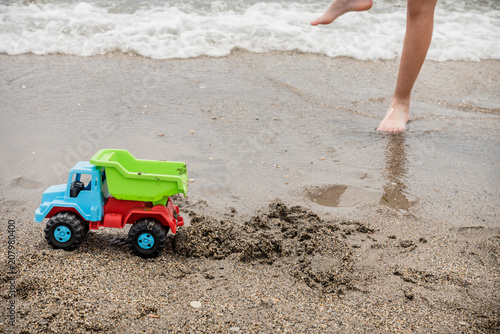 green, blue and red toy truck on the seashore in front of a child's feet