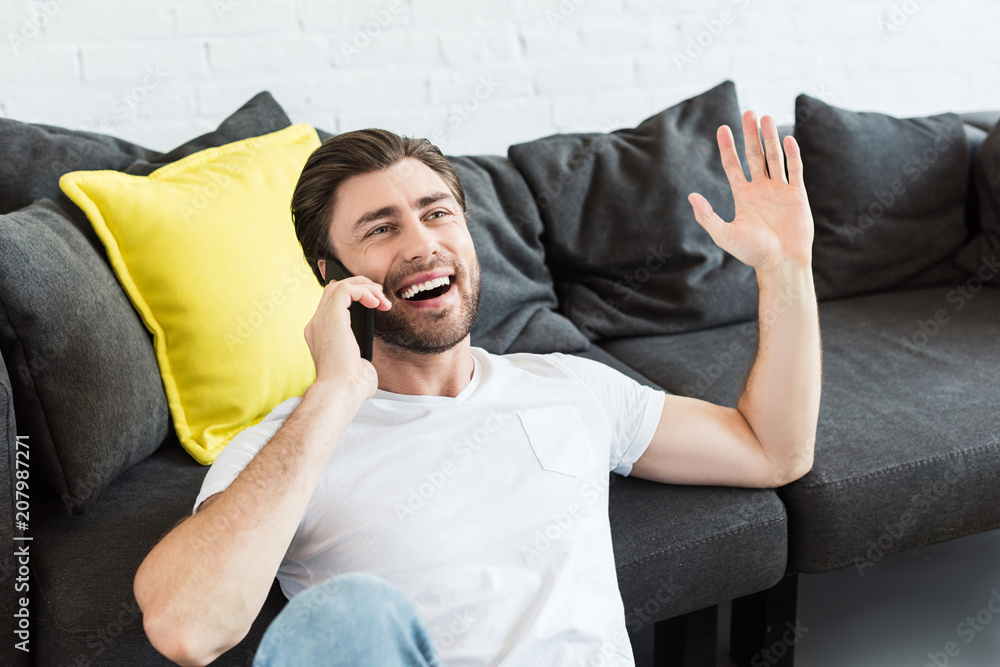 Fototapeta premium young man gesturing by hand and talking on smartphone near couch at home