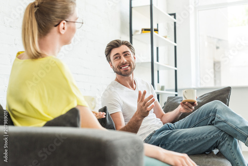 young smiling man with coffee cup gesturing by hand and talking to girlfriend at home