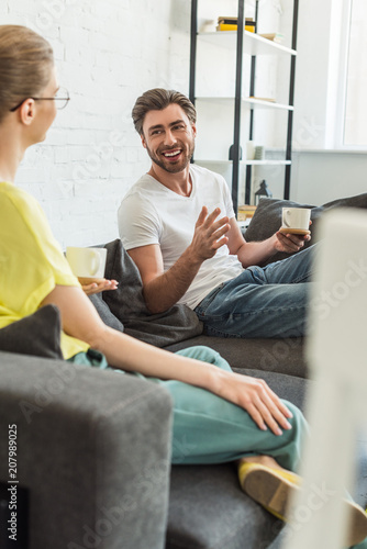 young smiling couple with coffee cups talking and sitting on sofa at home