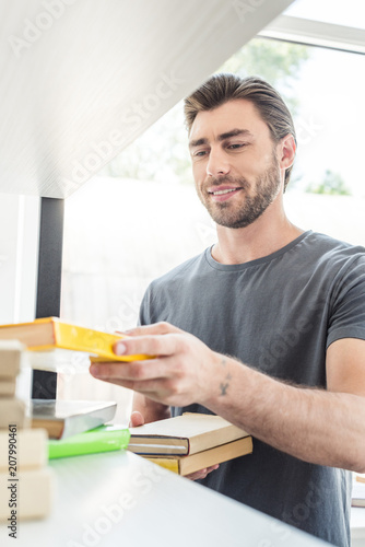young smiling man putting books on shelves at home