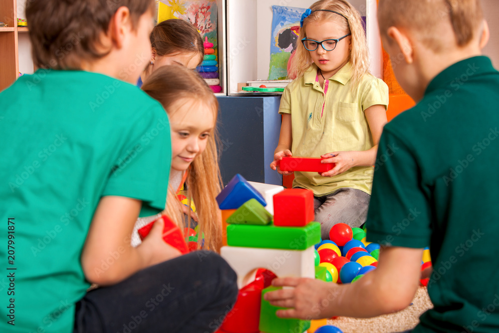 Fototapeta premium Children building blocks in kindergarten. Group kids playing toy on floor. Top view of interior preschool. Mat is big construction site building tower of cubes. Tournament of children's creativity.