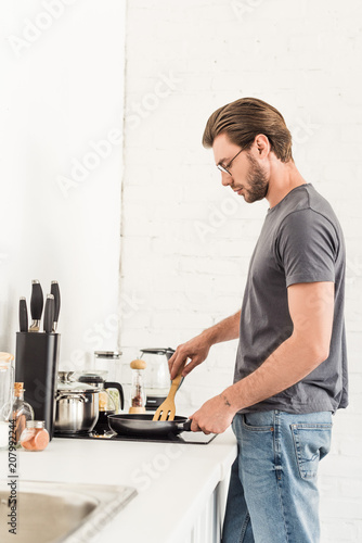 side view of young man cooking on stove with frying pan and spatula at kitchen