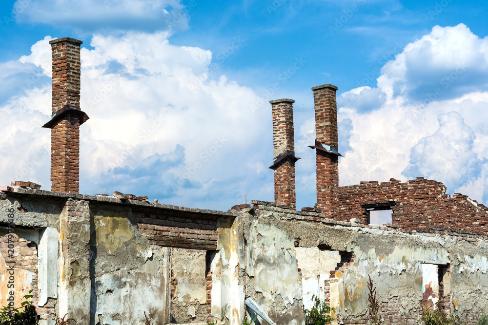 Abandoned destroyed old family brick house without roof and with chimneys, broken windows, window frames and bricks surrounded with grass on cloudy weather day.