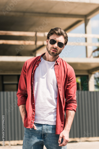 portrait of young stylish man in sunglasses posing in front of construction building