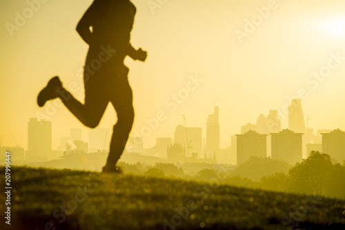 Photography Defocused silhouette of jogger running on a green grassy hill in front of the misty London city skyline