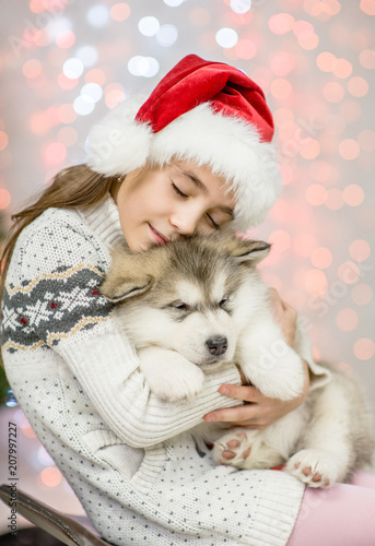  little girl in red santa hat hugging puppy on a background of the Christmas tree
