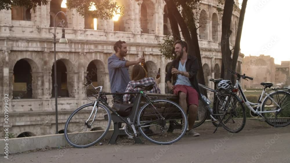 Three young friends tourists with bikes sitting on bench in front of colosseum under tree at sunset drinking beers having fun talking laughing chilling in Rome slow motion Three young friends tourists with bikes sitting on bench in front of colosseum under tree at sunset drinking beers having fun talking laughing chilling in Rome slow motion