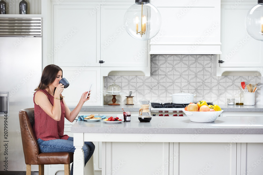 Woman reading the news on her phone in the morning before work in kitchen Woman reading the news on her phone in the morning before work in kitchen